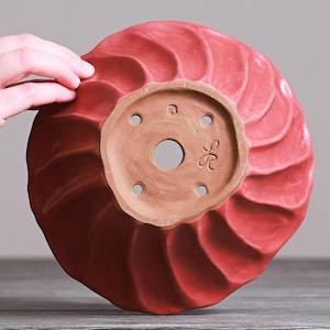 May include: A circular red ceramic bonsai pot featuring a spiraling, fluted texture on the exterior. The unglazed terracotta base includes a large central drainage hole, four smaller surrounding holes, and two distinct maker's marks stamped into clay.