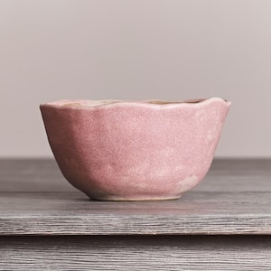 May include: A small, round ceramic bowl with a dusty pink, textured matte finish. The bowl features an irregular, wavy rim and a slightly uneven surface, giving it a hand-crafted appearance. The interior and exterior share the same pink tone.