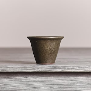 May include: A small, tapered ceramic cup with a dark, mottled olive-green glaze and a smooth, flared rim. The base of the vessel shows a thin, reddish-brown clay ring, contrasting with the textured, earthy finish of the exterior surface.