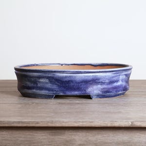 May include: A blue ceramic bonsai pot with a wide, oval shape. The pot has a textured, glazed finish with shades of blue and purple. The interior is a natural terracotta color. The pot sits on a wooden surface.