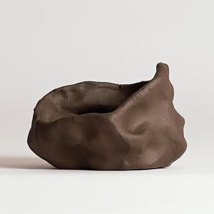 May include: A handmade, dark brown ceramic bowl with an organic, textured shape. The bowl has a unique, asymmetrical design with a raised edge on one side. The interior is a deep, dark space, perfect for holding small items or as a decorative piece.