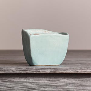 May include: A small, light blue ceramic planter with an irregular, organic shape and a smooth, matte finish. The vessel features a wide, open top with slightly wavy, uneven edges and a flat base, showcasing a simple, hand-crafted appearance.