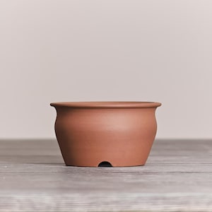 May include: A terracotta-colored ceramic pot with a smooth, matte finish. The vessel features a wide, rounded body that tapers toward a flat base, which includes a small, semi-circular drainage cutout at the bottom edge and a flared rim.