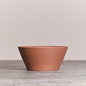 May include: A terracotta-colored bowl with a smooth, matte finish and a wide, flared rim. The bowl features a simple, tapered design with a flat base and subtle, light-colored speckles distributed across the exterior surface of the clay.