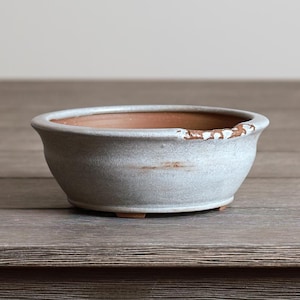 May include: A small, round, gray ceramic bonsai pot with a distressed rim. The pot has a light gray exterior and a reddish-brown interior. It sits on three small feet and is suitable for small plants.