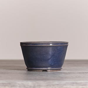 May include: A deep blue ceramic bowl with a smooth, glossy finish and a slightly flared rim. The bowl features a subtle gradient of darker blue tones and a defined base, showcasing a simple, functional design with a clean, rounded silhouette.