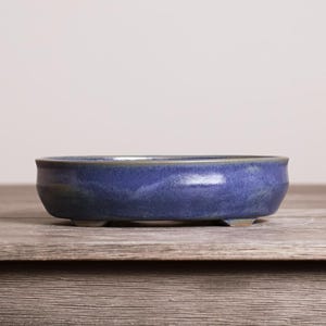 May include: A shallow, oval-shaped ceramic bonsai pot with a deep blue glaze featuring subtle variations in tone. The vessel has a smooth, rounded rim and is supported by small, integrated feet at the base, presenting a clean and functional design.