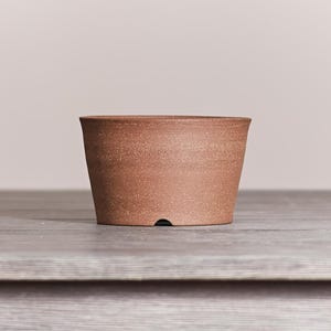 May include: A terracotta-colored ceramic planter with a wide, open top and a tapered body. The surface has a matte, speckled texture throughout. A small, semi-circular drainage notch is visible at the base of the planter on the front side.