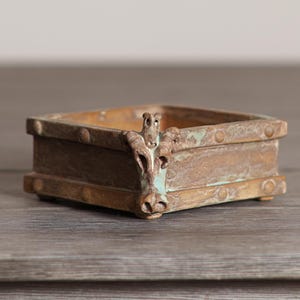 May include: A square, antique-style trinket box with a weathered, bronze-colored finish. The box features a decorative animal head on one corner and rivet details. The interior appears to be a lighter shade of brown.