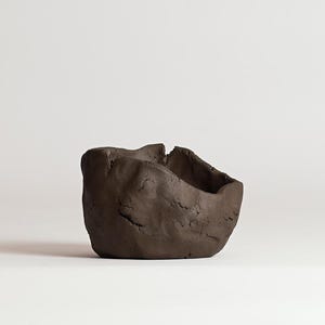 May include: A handmade, dark brown clay bowl with a rough, textured surface. The bowl has an irregular shape with a wide opening and a slightly uneven rim. The interior is smooth, contrasting with the exterior's rugged appearance. The bowl is likely for decorative use.