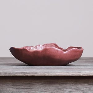 May include: A shallow, hand-formed ceramic bowl with a matte, dusty rose finish. The bowl features an irregular, organic, and wavy rim that mimics natural, flowing shapes, creating a unique silhouette with a smooth, consistent surface texture.