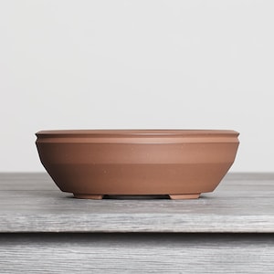 May include: A shallow, round terracotta bonsai pot with a matte finish. The pot features a wide, flat rim and a tapered body that narrows toward the base, which is supported by small, integrated feet. The surface is smooth and uniform in color.