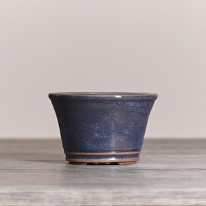 May include: A small, tapered ceramic bonsai pot with a deep blue, mottled glaze finish. The pot features a smooth, flared rim and a visible unglazed brown clay base at the bottom, showcasing a simple, functional design for small plant cultivation.