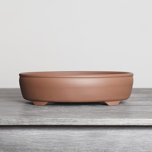 May include: A shallow, round, terracotta-colored ceramic bonsai pot with a smooth matte finish. The vessel features a slightly curved wall, a flat rim, and is supported by small, integrated feet at the base, presenting a simple, functional design.