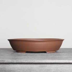 May include: A shallow, oval-shaped terracotta bonsai pot with a smooth, matte finish. The vessel features a wide, open top with a slightly flared rim and is supported by small, integrated feet at the base, showcasing a natural reddish-brown clay color.