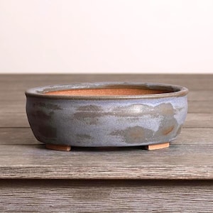 May include: A small, oval-shaped ceramic bonsai pot. The pot has a blue-gray exterior with brown accents and a reddish-brown interior. It sits on three small, rectangular feet. The pot is designed for planting small trees or plants.