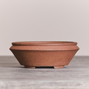 May include: A shallow, round terracotta bonsai pot with a speckled, matte finish. The vessel features a wide, angled rim that tapers down to a narrow base supported by small, integrated feet. The clay is a warm, earthy reddish-brown color.