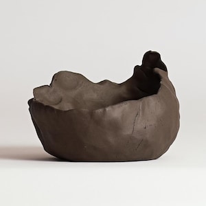 May include: A handmade, dark brown ceramic bowl with an organic, textured shape. The bowl has a wide, irregular rim and a rough, unglazed surface. The interior is smooth, contrasting with the exterior. The bowl is likely used for decorative or functional purposes.