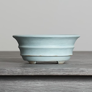 May include: A light blue ceramic bonsai pot with a flared rim and a tiered, ribbed exterior design. The vessel features a wide, open top and is supported by three small, sturdy feet at the base, showcasing a smooth, matte finish throughout.