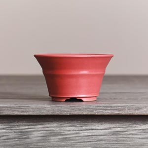 May include: A matte red ceramic bonsai pot with a wide, flared rim and a tapered body. The vessel features a smooth, solid-colored finish and a raised base, showcasing a clean, functional design suitable for small plant cultivation and display.