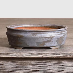 May include: A handcrafted, oval-shaped bonsai pot. The ceramic pot features a weathered, gray-blue exterior with a reddish-brown interior. The pot is supported by three small feet and sits on a wooden surface.