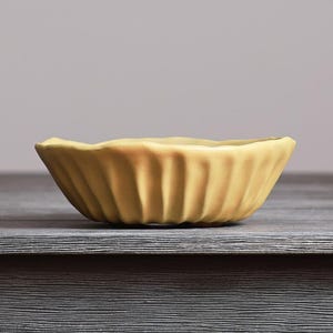 May include: A small, round, yellow ceramic bowl with a fluted, scalloped exterior design. The bowl features a matte finish and a wavy, uneven rim, creating a textured appearance around the sides of the vessel, which is presented in a side profile view.
