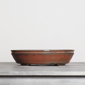 May include: A shallow, oval-shaped ceramic bonsai pot with a rustic terracotta-colored finish. The vessel features a dark, weathered glaze along the rim and base, supported by four small, integrated feet that elevate the structure slightly.