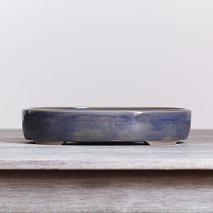 May include: A shallow, oval-shaped ceramic bonsai pot with a glossy, deep blue glaze that transitions into subtle earthy brown tones along the base. The pot features a smooth, rounded rim and is supported by small, integrated feet at the bottom.