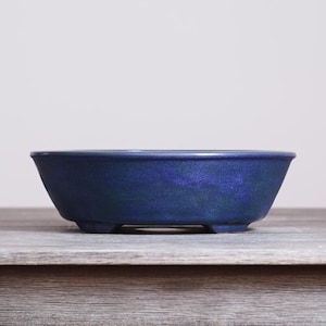May include: A deep blue ceramic bonsai pot with a wide, oval-shaped opening and a smooth, slightly mottled finish. The vessel features a thick, rounded rim and is supported by four small, sturdy feet at the base, providing a stable foundation.