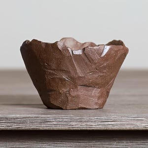 May include: A small, hand-formed ceramic bowl with a dark brown, earthy tone. The exterior features a rugged, faceted texture with sharp, angular indentations and a rough, uneven rim, giving it a raw, stone-like appearance and a matte finish.