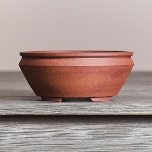 May include: A terracotta-colored ceramic bonsai pot with a wide, circular opening and a distinct angled rim. The bowl features a matte, earthy texture with a tapered body that rests on small, integrated feet, showcasing a simple, functional design.