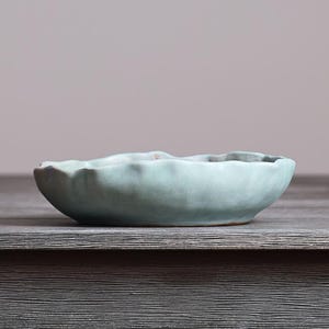 May include: A shallow, round ceramic bowl with a matte, light blue-green glaze. The bowl features an organic, uneven, and wavy rim that creates a textured, hand-formed appearance. The surface of the bowl is smooth with subtle color variations.