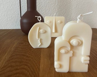 May include: Three white ceramic candles shaped like abstract faces. The candles are on a wooden surface next to a brown ceramic vase.