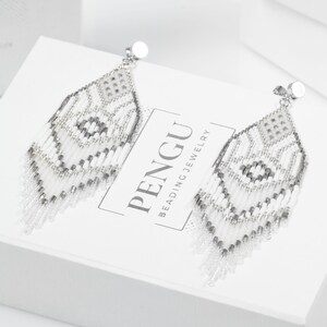 May include: White and silver beaded dangle earrings with a geometric pattern. The earrings are on a white box with the text "PENGU BEADING JEWELRY".