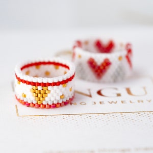 May include: Two beaded rings with geometric patterns. One ring is white with red and gold accents, and the other is white with red accents. The rings are on a white surface with gold lettering that reads "NGU JEWELRY".