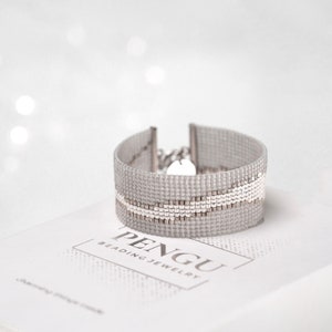 K&ouml;nnte beinhalten: Ein silbernes Armband mit einem wei&szlig;en geometrischen Muster. Das Armband liegt auf einer wei&szlig;en Schachtel mit dem Text "PENGU BEADING JEWELRY".