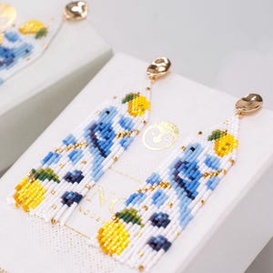 Puede incluir: Pendientes blancos con cuentas con un diseño floral con flores azules y limones amarillos. Los pendientes tienen tachuelas doradas y pequeñas cuentas doradas. Los pendientes se exhiben sobre una superficie blanca.