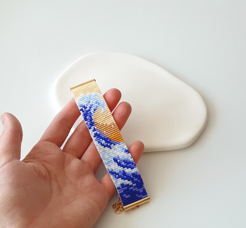K&ouml;nnte beinhalten: Ein Armband aus Perlen mit einem blauen und gelben Wellendesign. Das Armband ist aus kleinen Perlen gefertigt und hat einen goldenen Verschluss.