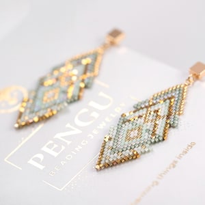 May include: A pair of green and gold beaded earrings with a geometric design. The earrings are hanging from gold hooks.