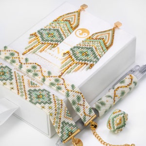 Può includere: Un set di gioielli in perle verdi e dorate, tra cui un paio di orecchini, due braccialetti, un anello e una penna con perle. I gioielli sono esposti in una scatola bianca con la scritta "charming things inside".