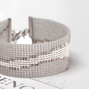 K&ouml;nnte beinhalten: Ein silbernes und wei&szlig;es Perlenarmband mit einem wellenf&ouml;rmigen Muster. Das Armband besteht aus kleinen, quadratischen Perlen und hat einen silbernen Verschluss.