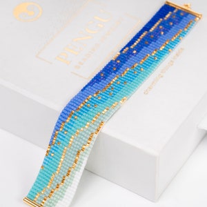 Peut inclure: Un bracelet en perles bleu et or avec un dégradé de couleurs, du bleu clair au bleu foncé. Le bracelet est sur une boîte blanche avec le mot "PENGU" imprimé dessus.