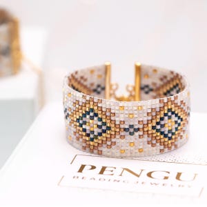Può includere: Bracciale largo con perline con un motivo geometrico bianco, oro, blu navy e bronzo. Il bracciale ha una chiusura dorata ed è esposto su una superficie bianca. Il marchio "PENGU BEADING JEWELRY" è visibile.