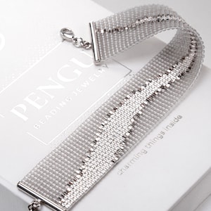 Può includere: Un braccialetto di perline argentate con una chiusura argentata. Il braccialetto è su una scatola bianca con il testo "PENGU BEADING JEWELRY" e "charming things inside".