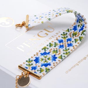 Peut inclure: Un bracelet de perles blanc avec un motif géométrique bleu et vert. Le bracelet a des fermoirs dorés et un petit charme. Le texte "PENGU BEADING JEWELRY" est visible en arrière-plan.