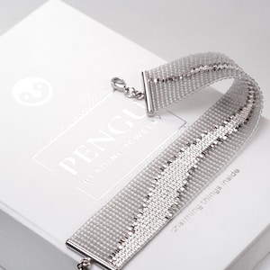 K&ouml;nnte beinhalten: Ein silbernes Perlenarmband mit einem silbernen Verschluss. Das Armband liegt auf einer wei&szlig;en Schachtel mit dem Text "PENGU Beading Jewelry" und "charming things inside".