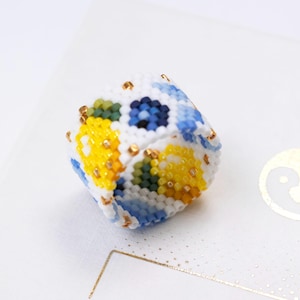 Può includere: Oggetto cubico fatto a mano con perline. Il lavoro con le perline presenta una base bianca con motivi floreali gialli, blu e verdi. Perline color oro accentuano i bordi. L'oggetto è probabilmente un oggetto decorativo o un gioiello unico.
