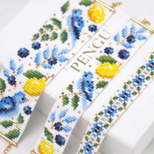 Könnte beinhalten: Nahaufnahme mehrerer Perlenarmbänder mit floralen Designs. Die Armbänder haben weiße Hintergründe mit blauen Blumen, gelben Zitronen, grünen Blättern und goldenen Akzenten. Die Armbänder werden auf einer weißen Oberfläche präsentiert.