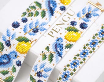May include: Close-up of several beaded bracelets with floral designs. The bracelets feature white backgrounds with blue flowers, yellow lemons, green leaves, and gold accents. The bracelets are displayed on a white surface.