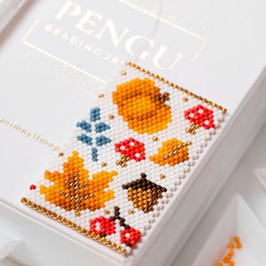 Puede incluir: Un diseño otoñal con cuentas que presenta una calabaza, una hoja de arce, setas y una bellota. El diseño es blanco con bordes y detalles dorados, y los elementos otoñales son naranjas, rojos, marrones y azules. Sobre una caja blanca.